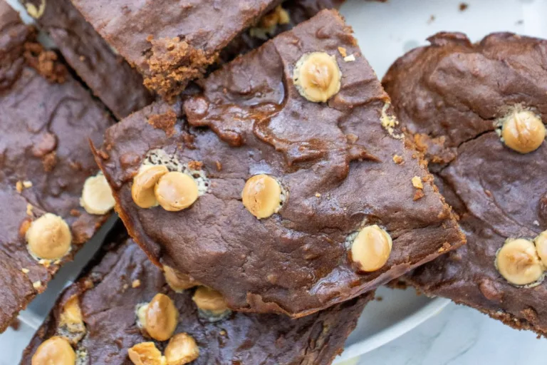 Close-up of several Sweet Potato Brownies topped with scattered white chocolate chips, stacked on a white plate. The brownies appear rich and fudgy with a slightly cracked surface.