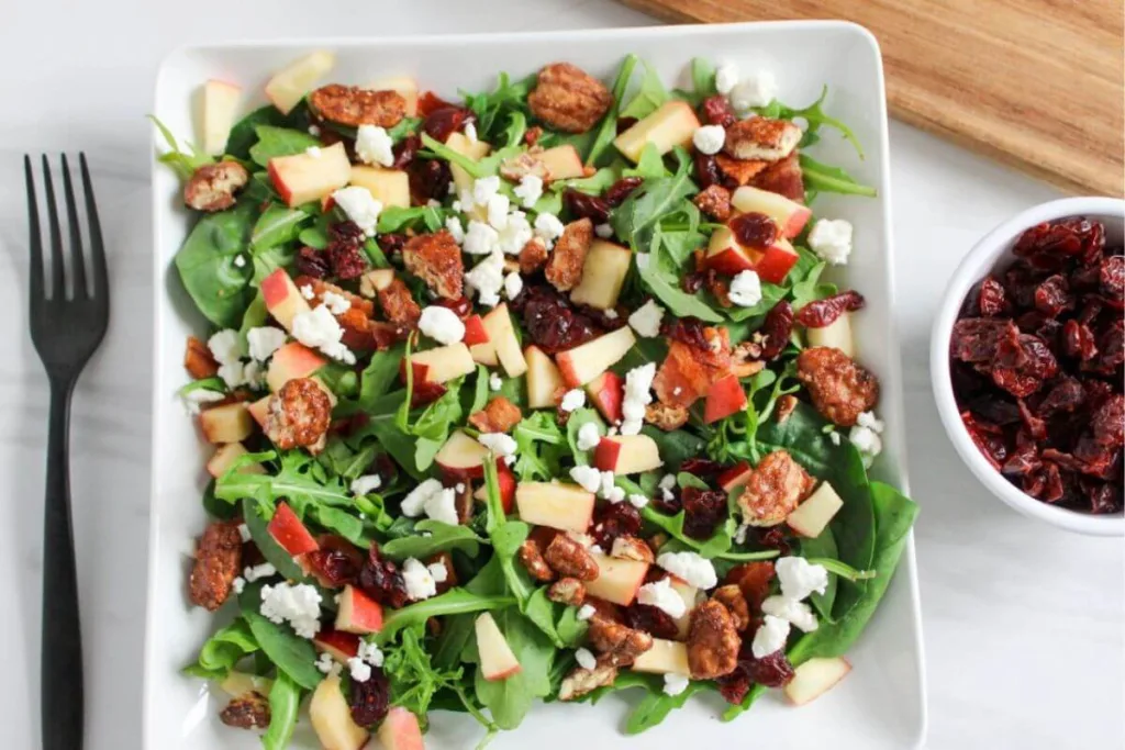 A square white plate with a fresh salad of spinach, arugula, diced apples, candied pecans, dried cranberries, and crumbled goat cheese. A fork and a small bowl of dried cranberries are beside the plate.