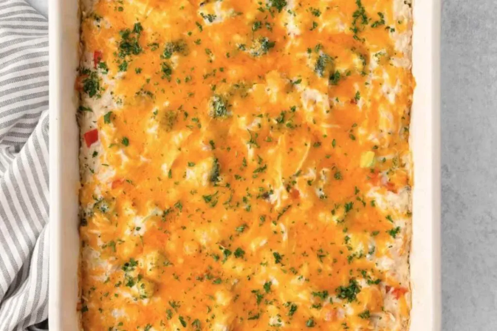 A baked casserole in a white dish, topped with melted cheese and sprinkled with chopped herbs. The surface is golden and slightly bubbly, with visible bits of vegetables underneath. A striped kitchen towel is partially visible on the side.