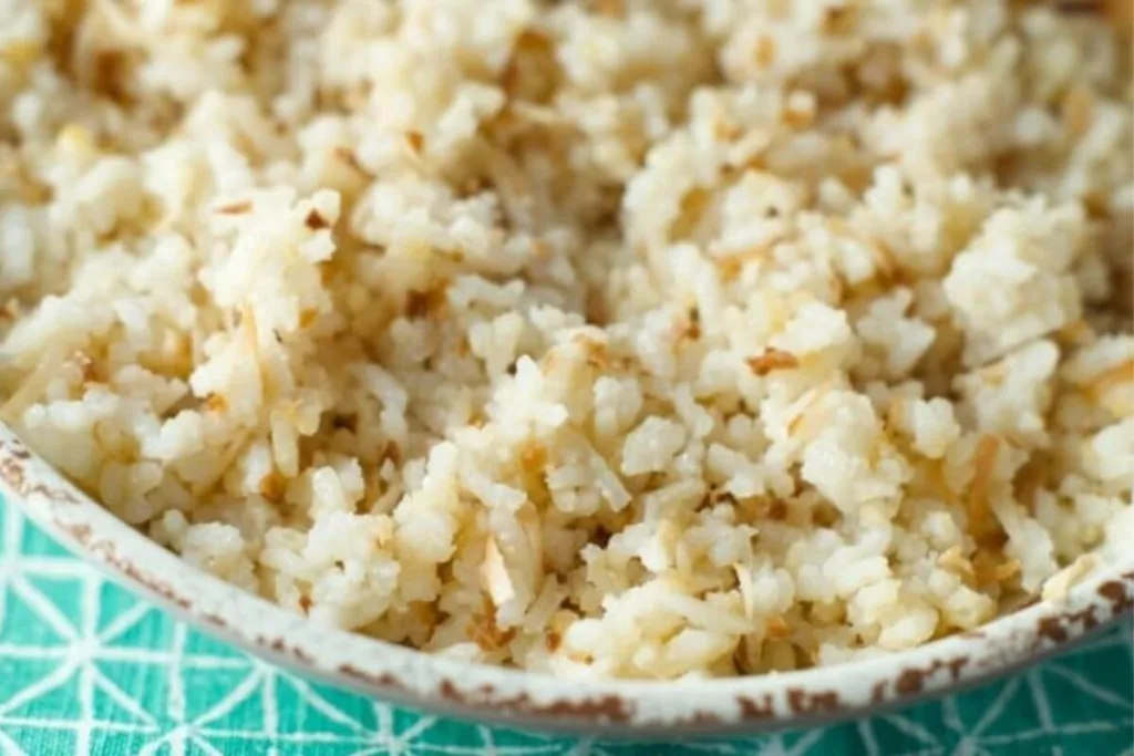 A close-up of a bowl filled with lightly toasted coconut rice, showing fluffy white rice mixed with shredded coconut on a patterned green and white surface.