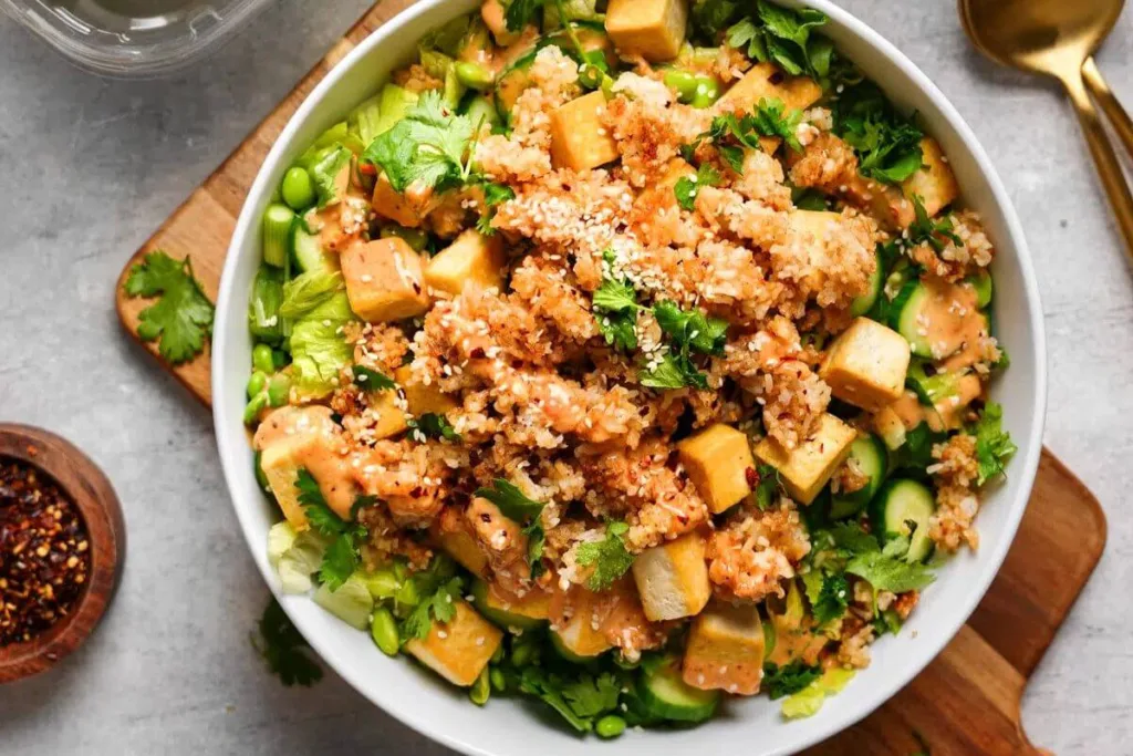 A bowl of salad with cubes of tofu, quinoa, sliced cucumbers, edamame, fresh cilantro, and a creamy orange dressing, garnished with sesame seeds&mdash;perfect alongside budget friendly rice meals. Utensils and red pepper flakes are served nearby.