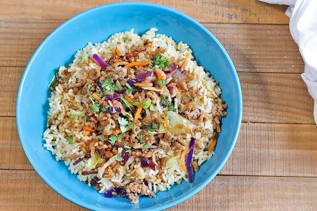 A blue bowl filled with cooked rice topped with ground meat, shredded carrots, cabbage, purple cabbage, and fresh herbs sits on a wooden table&mdash;perfect for those seeking delicious budget friendly rice meals.