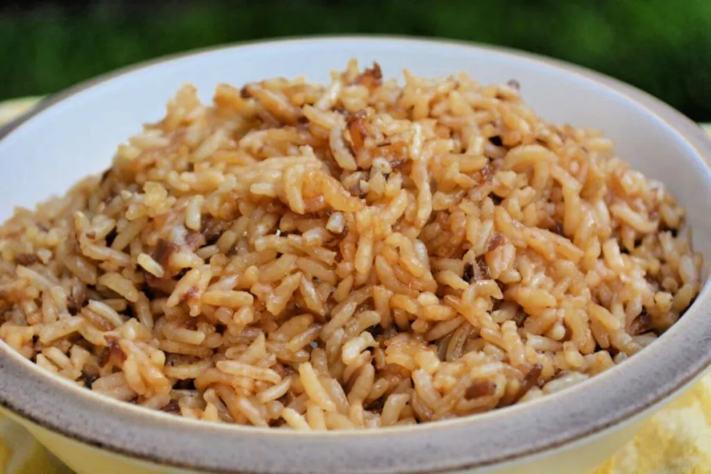 A bowl filled with cooked, seasoned rice that appears brown and slightly glossy, suggesting it&rsquo;s a Budget Friendly Rice Meal mixed with soy sauce or seasonings. The green background is softly out of focus.