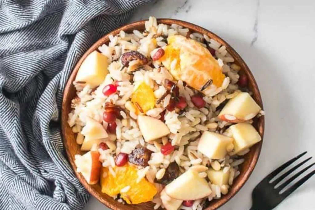 A wooden bowl filled with rice salad containing chopped apples, orange segments, pomegranate seeds, and dates, placed on a marble surface beside a gray-striped cloth and a black fork.