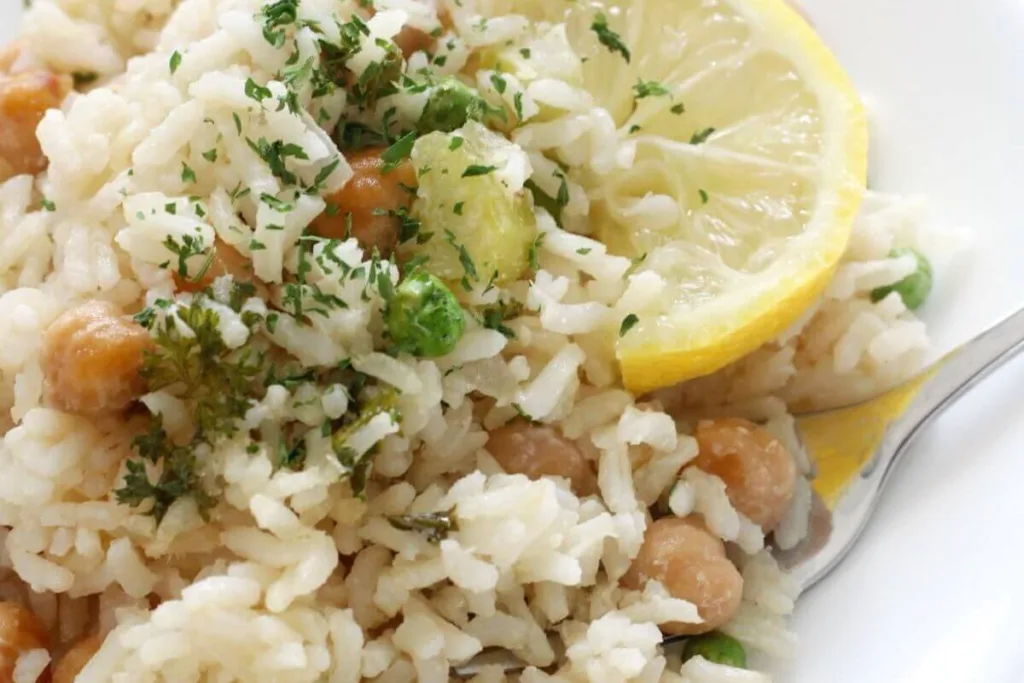 A close-up of cooked rice mixed with chickpeas and green peas, garnished with chopped herbs and two lemon slices, with a silver fork beside the food on a white plate.