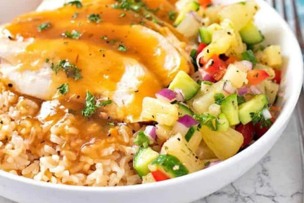 A bowl of brown rice topped with chicken and golden sauce, served with a colorful side of pineapple salsa containing cucumber, red onion, tomato, herbs, and black pepper.
