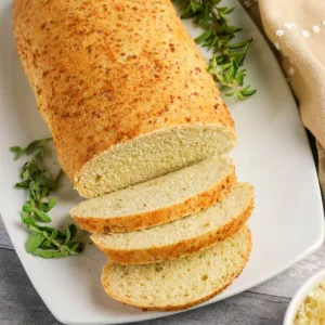 Parmesan Oregano Bread sliced sitting on a white plan with fresh herbs around it.