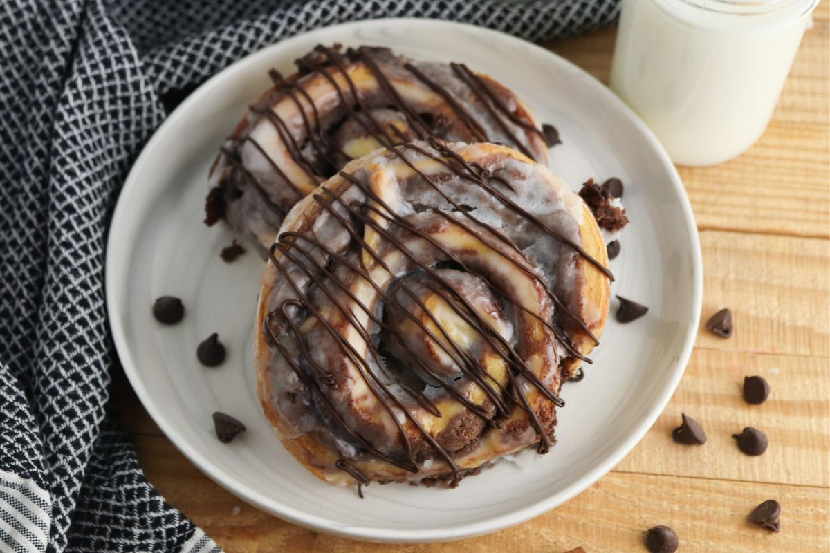 Cinnamon rolls with brownie mixture sitting on a white plate with chocolate chips and a glass of milk.